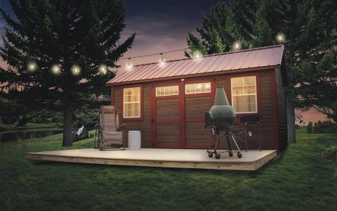 Tiny house made from a garden shed with brown siding, red trim, and red roofing sitting by a river with a porch that has hanging lights, a grill, and outdoor seating.